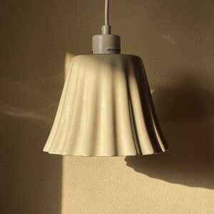 May include: White ceramic lampshade with a ruffled design. The lampshade is hanging from a white cord and has the word "androw" printed on the bottom.