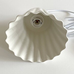 May include: White ceramic lampshade with a ruffled edge. The lampshade has a standard light bulb socket.