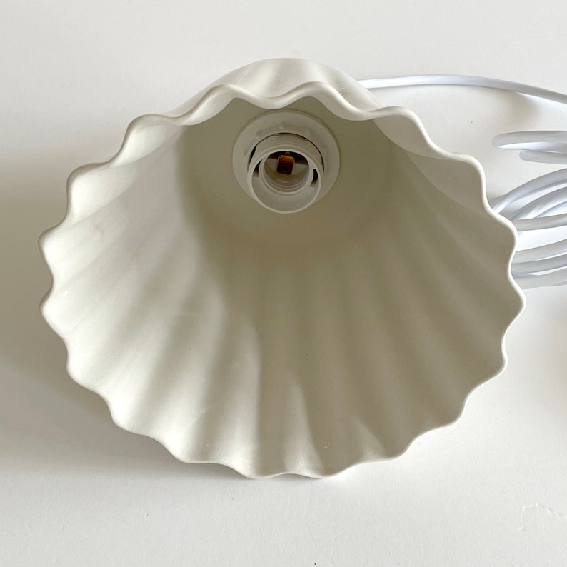 May include: White ceramic lampshade with a ruffled edge. The lampshade has a standard light bulb socket.