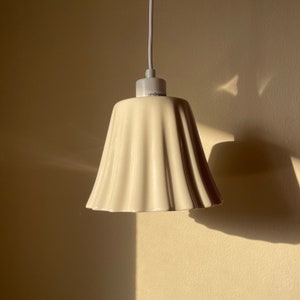 May include: A white ceramic pendant light fixture with a ruffled shade. The light fixture is hanging from a white cord and has a white ceiling mount.