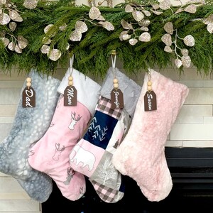 May include: Four Christmas stockings hang from a fireplace mantel, each with a wooden tag. The stockings are gray, pink, and patterned with deer, trees, and a bear. The tags read "Dad", "Mom", "Jennifer", and "Susan".