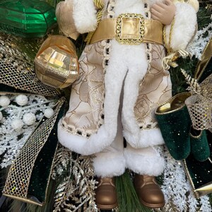 May include: A festive Christmas wreath featuring a Santa Claus figurine. The Santa figure is dressed in a cream-colored coat with gold accents and white faux fur trim. The wreath includes green, gold, and white decorative elements.