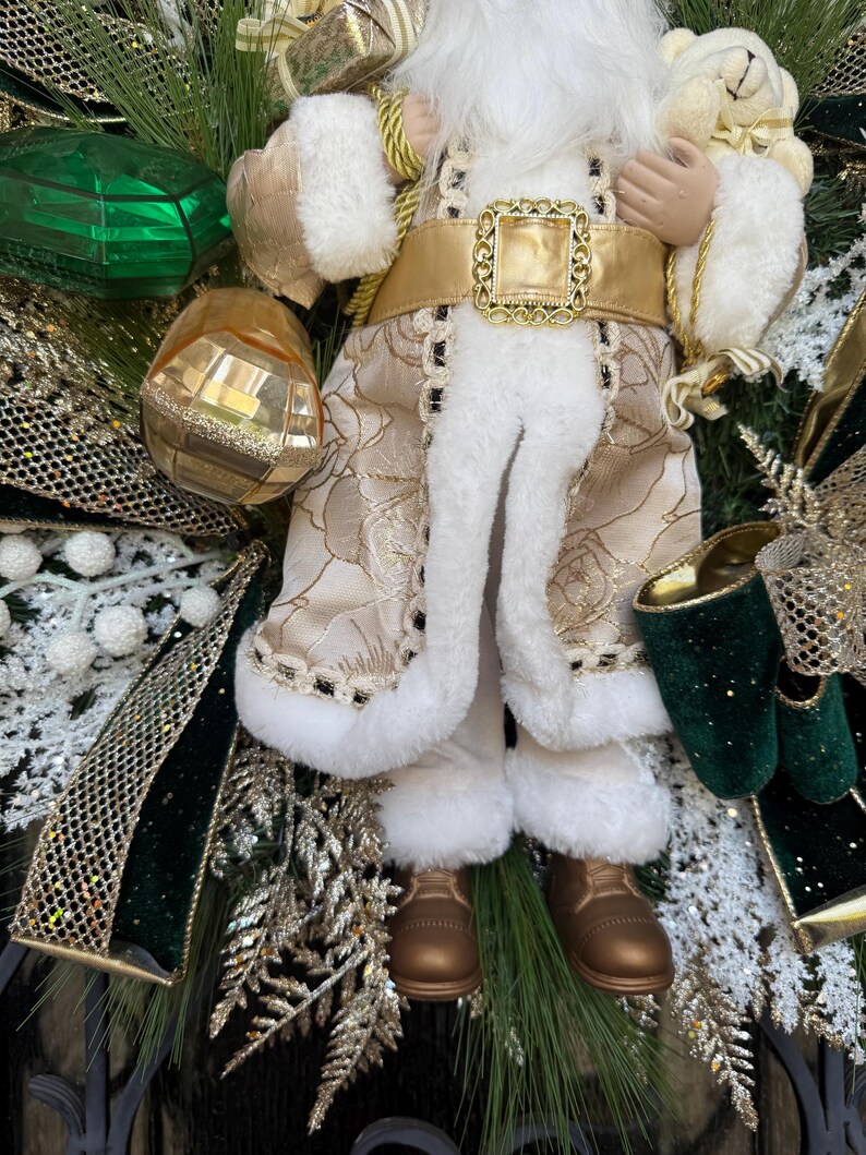 May include: A festive Christmas wreath featuring a Santa Claus figurine. The Santa figure is dressed in a cream-colored coat with gold accents and white faux fur trim. The wreath includes green, gold, and white decorative elements.