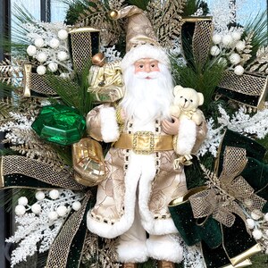 May include: A festive Christmas wreath featuring a Santa Claus figurine holding a teddy bear. The wreath includes green pine branches, white berries, gold ribbons, and a large green gem. The Santa Claus figure is dressed in a gold and white coat.
