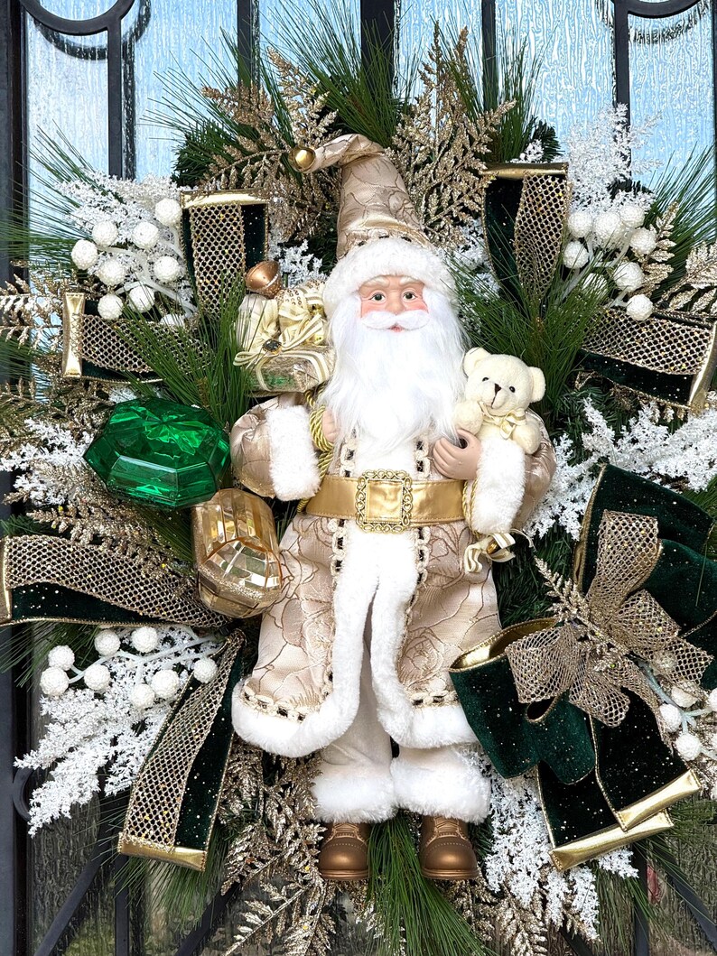 May include: A festive Christmas wreath featuring a Santa Claus figurine holding a teddy bear. The wreath includes green pine branches, white berries, gold ribbons, and a large green gem. The Santa Claus figure is dressed in a gold and white coat.