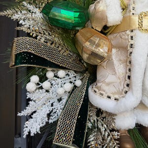 May include: A festive Christmas wreath featuring a Santa Claus figure, green and gold ornaments, and a decorative bow. The wreath includes faux evergreen branches, white berries, and gold glitter accents. The Santa Claus figure is wearing a white and gold outfit.