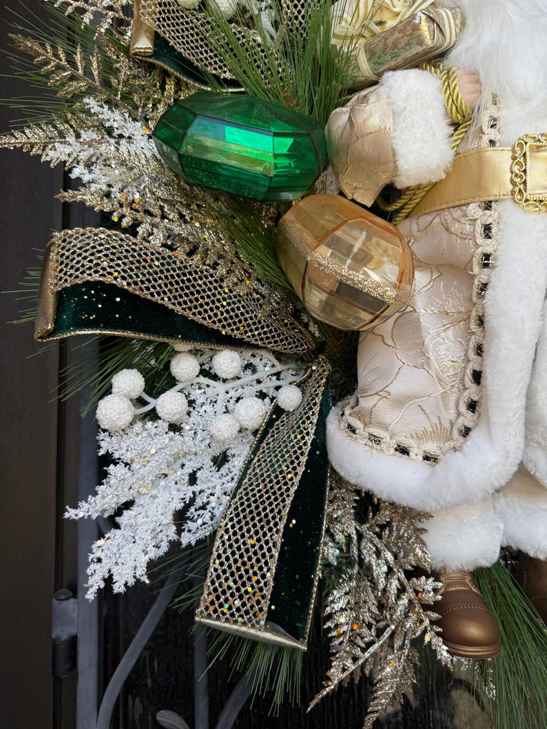 May include: A festive Christmas wreath featuring a Santa Claus figure, green and gold ornaments, and a decorative bow. The wreath includes faux evergreen branches, white berries, and gold glitter accents. The Santa Claus figure is wearing a white and gold outfit.