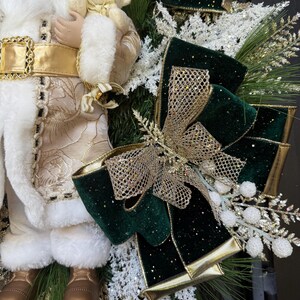 May include: A festive Christmas decoration featuring a Santa Claus figurine holding a teddy bear. The Santa is dressed in a white and gold outfit with a gold belt. A green and gold bow and decorative greenery complete the holiday theme.