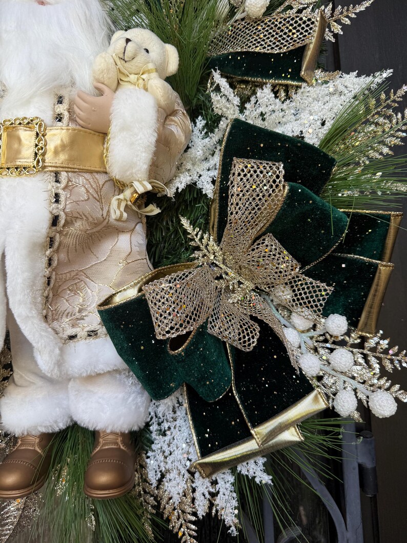May include: A festive Christmas decoration featuring a Santa Claus figurine holding a teddy bear. The Santa is dressed in a white and gold outfit with a gold belt. A green and gold bow and decorative greenery complete the holiday theme.