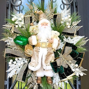 May include: A festive Christmas wreath featuring a Santa Claus figurine holding a teddy bear and gifts. The wreath is adorned with gold and green ribbons, white accents, and faux greenery, creating a holiday display.