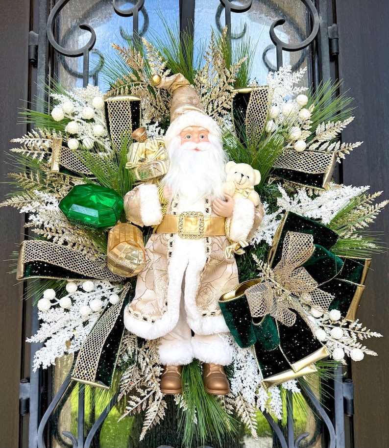 May include: A festive Christmas wreath featuring a Santa Claus figurine holding a teddy bear and gifts. The wreath is adorned with gold and green ribbons, white accents, and faux greenery, creating a holiday display.