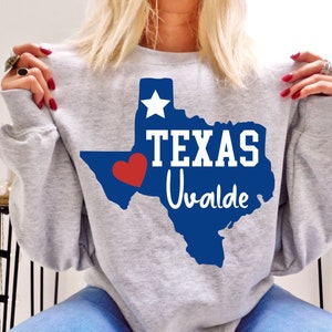Peut inclure: Un sweat-shirt gris clair avec un contour bleu de l'État du Texas, une étoile blanche et le mot "TEXAS" en blanc. En dessous, "Uvalde" est en écriture blanche. Un cœur rouge est sur le côté inférieur gauche du Texas. Vêtement décontracté.