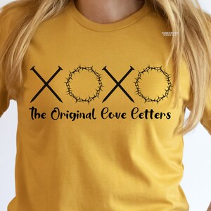 May include: Mustard yellow t-shirt with a black graphic design. The design includes two crossed nails, a crown of thorns, and the text "The Original Love Letters". The shirt is a crew neck style.