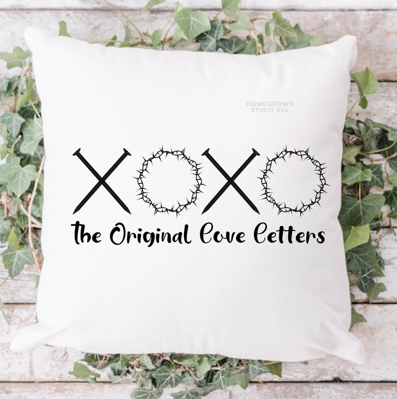 May include: White square pillow with black graphic text and symbols. The design includes two crossed nails, a crown of thorns, and the words "The Original Love Letters". The pillow is surrounded by green leaves.