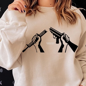 May include: Beige sweatshirt with a black graphic of two hands breaking a rifle in half. The text "HOMEGROWN STUDIO USA" is printed below the graphic. The sweatshirt has long sleeves and a crew neck.