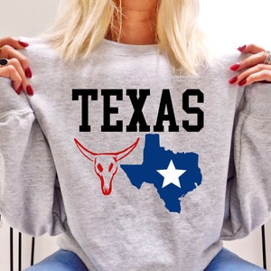 Peut inclure: Un sweat-shirt gris clair avec le mot "TEXAS" en noir gras. En dessous, un crâne de longhorn rouge et une carte bleue du Texas avec une étoile blanche.