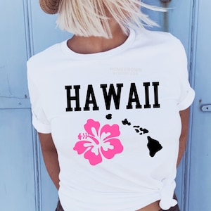 Peut inclure: T-shirt blanc avec le mot "HAWAII" en noir gras, une fleur d'hibiscus rose et une silhouette noire des îles hawaïennes. Le t-shirt porte également le texte "HOMEGROWN STUDIO USA".