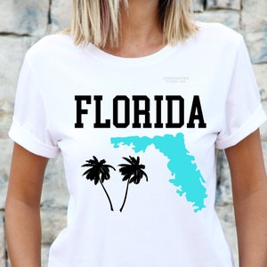 Peut inclure: T-shirt blanc avec le mot "FLORIDA" en lettres noires, un contour turquoise de l'État et deux palmiers noirs. Le t-shirt a des manches retroussées et un col rond.