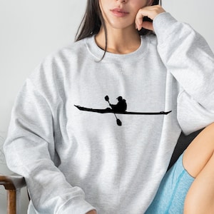 Pode incluir: Camisola cinza claro com uma silhueta preta de um caiaquista. O texto "HOMEBURG TO THE SEA" está impresso em branco no topo. A camisola tem mangas compridas e gola redonda.