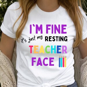 K&ouml;nnte beinhalten: Wei&szlig;es T-Shirt mit dem Text "I'm Fine it's just my resting teacher face" in Lila, Schwarz und Regenbogenfarben. Vier bunte Stifte befinden sich rechts neben dem Text.