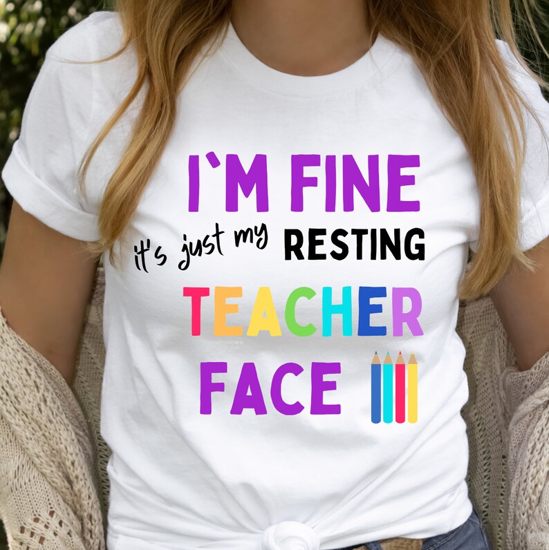 K&ouml;nnte beinhalten: Wei&szlig;es T-Shirt mit dem Text "I'm Fine it's just my resting teacher face" in Lila, Schwarz und Regenbogenfarben. Vier bunte Stifte befinden sich rechts neben dem Text.