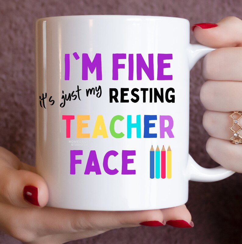 K&ouml;nnte beinhalten: Wei&szlig;e Keramiktasse mit dem Text "I'm fine it's just my resting teacher face" in Lila, Schwarz und Regenbogenfarben. Vier bunte Bleistifte befinden sich rechts neben dem Text.