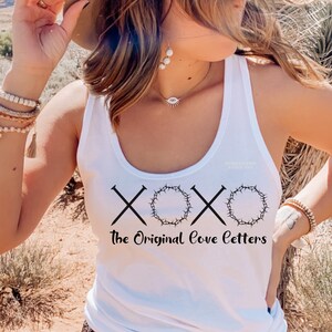 May include: White tank top with a black graphic design. The design includes the letters "XOX" with nail and crown of thorns imagery, and the text "The Original Love Letters". The tank top is sleeveless.