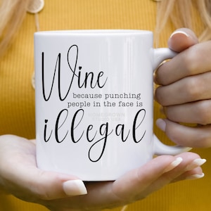 May include: White ceramic mug with the words "Wine because punching people in the face is illegal" in black script. The mug has a standard handle and is held by a person.