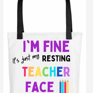 K&ouml;nnte beinhalten: Wei&szlig;e Einkaufstasche mit schwarzen Henkeln und dem Text "I'm fine it's just my resting teacher face" in bunten Buchstaben mit einer Reihe bunter Bleistifte darunter.