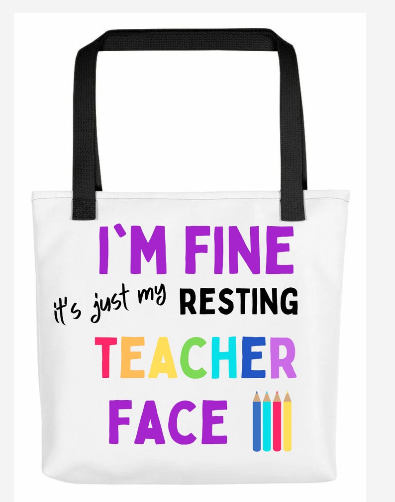 K&ouml;nnte beinhalten: Wei&szlig;e Einkaufstasche mit schwarzen Henkeln und dem Text "I'm fine it's just my resting teacher face" in bunten Buchstaben mit einer Reihe bunter Bleistifte darunter.