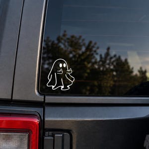 May include: A white vinyl decal sticker applied to a vehicle window, featuring a simple line-art illustration of a ghost with two round eyes, depicted with both arms raised and middle fingers extended in a gesture of defiance against a dark background.