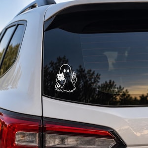 May include: A white vinyl decal applied to the rear window of a silver vehicle. The decal features a simple line drawing of a ghost holding a small cat in one arm and a tall iced beverage with a straw in the other hand, rendered in white lines.