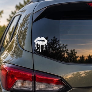May include: A white vinyl decal applied to the rear window of a dark-coloured vehicle. The decal features a graphic design of lips with sharp fangs, depicted with a dripping effect that extends downward from the lower lip in several vertical streams.