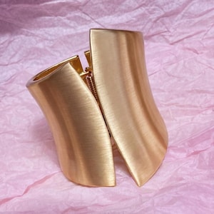 May include: A gold-toned cuff bracelet with a brushed metal finish. The bracelet has a curved, open design with a hinge and clasp closure. The jewelry is displayed on a pink, textured surface.
