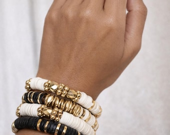 May include: A stack of bracelets in black, white, and gold. The bracelets are made of round, flat beads and gold-colored metal accents. The bracelets are stacked on a wrist.