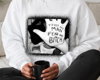 May include: A white long-sleeve sweatshirt featuring a black and white graphic print on the chest. The graphic depicts an open palm with the text "TRUST NO MAN FEAR NO BITCH" written across the fingers and palm, framed by a stippled black border.
