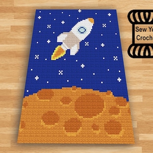 Puede incluir: Un patrón de crochet que presenta una nave espacial volando sobre un planeta con cráteres. El fondo es un cielo azul con estrellas blancas. El patrón se titula "Sew You Crochet".