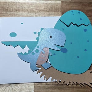 May include: A handmade greeting card featuring a light blue dinosaur emerging from a teal egg. The dinosaur has a light pink belly and is set against a white background with teal accents. The egg rests on a brown, textured base.