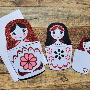May include: Three red and white nesting dolls with black and white details. The largest doll has a red glitter finish and a pink flower design. The middle doll has a black and white flower design. The smallest doll is plain white.