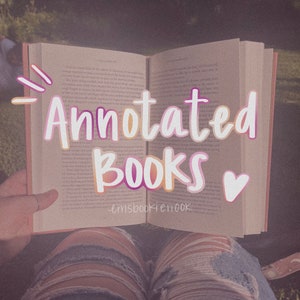 May include: A close-up of an open book with the text "Annotated Books" written in pink and yellow letters on the pages. The book is resting on a person's lap, and a pink heart is drawn on the right page.