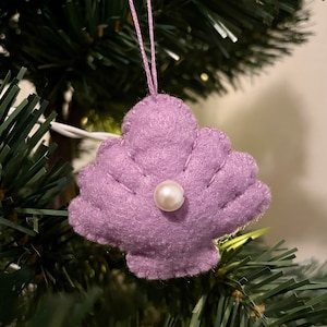May include: A purple felt ornament shaped like a seashell with a white pearl in the center. The ornament is hanging from a white string on a green Christmas tree.
