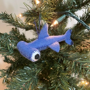 May include: A blue felt hammerhead shark ornament hanging on a green Christmas tree with white lights.