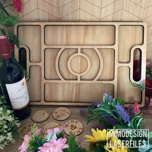 Op de afbeelding: Houten serveerblad met meerdere vakken, waaronder een ronde uitsparing voor een fles. Het blad heeft twee handvatten en is gemaakt van lichtbruin hout. Het blad is omringd door decoratieve onderzetters met zinnen als "Wine Down" en "Wine & Dine".