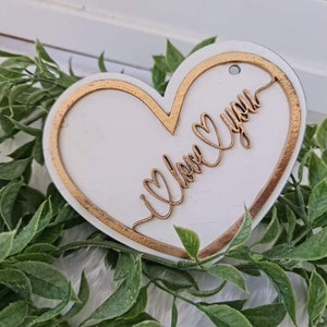 May include: A white heart-shaped wooden ornament with a gold border and the words "I love you" in gold script. The ornament has a small hole at the top for hanging. The ornament is surrounded by green foliage.