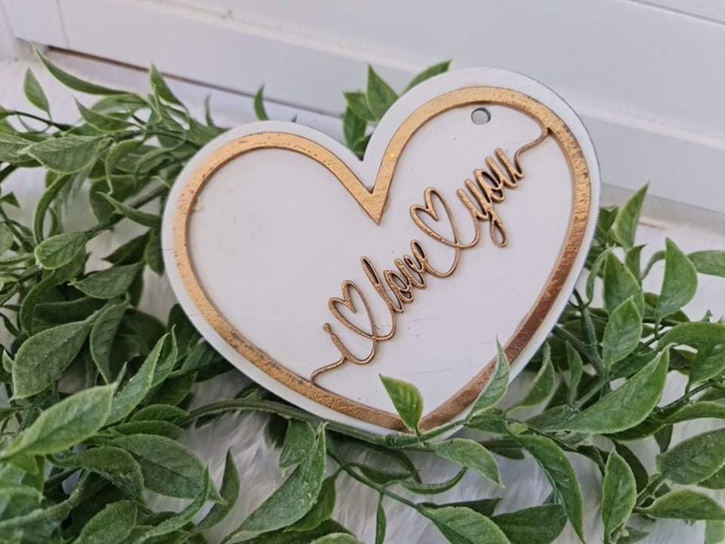 May include: A white heart-shaped wooden ornament with a gold border and the words "I love you" in gold script. The ornament has a small hole at the top for hanging. The ornament is surrounded by green foliage.