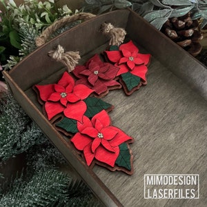 May include: Four red poinsettia flower ornaments with green leaves, arranged in a decorative pattern, on a wooden surface. The ornaments are laser cut and ready to be painted or stained. MIMODESIGN LASERFILES is printed on the surface.