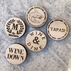 K&ouml;nnte beinhalten: F&uuml;nf runde Holz-Untersetzer mit lasergravierten Designs. Die Untersetzer zeigen den Text "Wine & Dine", "Wine Down", "The Mogensens", "Shark Coochie Board" und "Tapas!"
