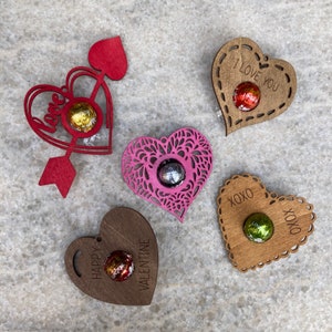 May include: Five wooden heart-shaped ornaments with different designs and sayings. Each ornament has a small round space to hold a chocolate. The sayings include "Love", "I Love You", "XOXO", "Happy Valentine", and a heart with a lace design.