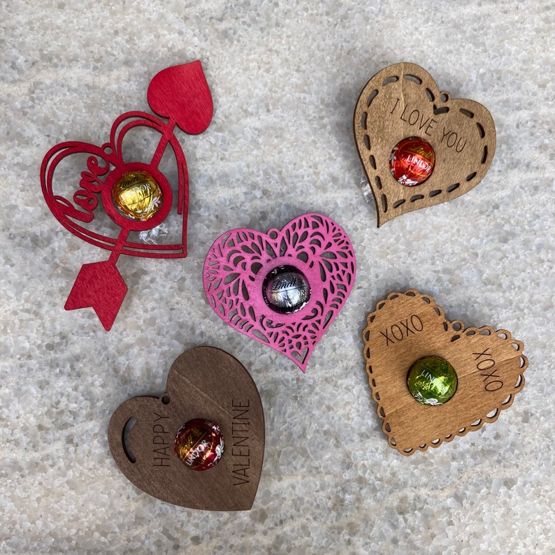 May include: Five wooden heart-shaped ornaments with different designs and sayings. Each ornament has a small round space to hold a chocolate. The sayings include "Love", "I Love You", "XOXO", "Happy Valentine", and a heart with a lace design.