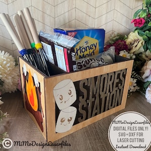 May include: Wooden box with the words "S'mores Station" on the front. The box is decorated with a campfire and marshmallows. The box is filled with graham crackers, chocolate bars, and marshmallows.